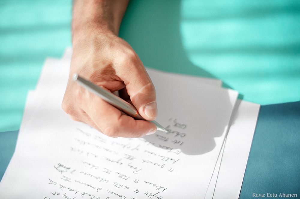 Kynää pitelevä käsi ja pöydällä pino papereita, joissa käsinkirjoitettua tekstiä.